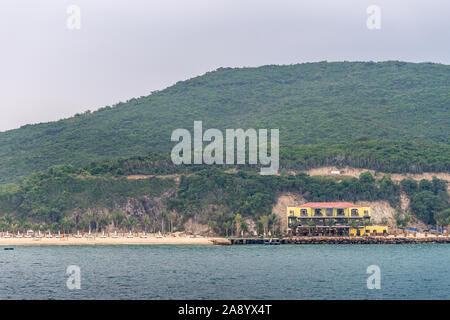 Nha Trang, Vietnam - Marzo 11, 2019: Serata, giallo con un tetto rosso beach house con i dock su Vinh Nguyen isola con collina boschiva nel retro. Giallo bea Foto Stock