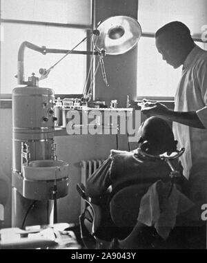 Vista interna di uno studio dentistico con un americano africano paziente seduto in poltrona e un dentista in piedi accanto a lui ca. 1947 Foto Stock