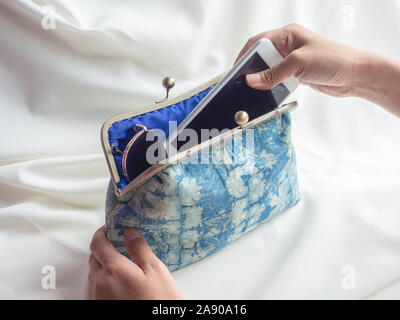 Donna mettendo mano smartphone bianco e occhiali da sole nel vintage blu indaco tessuto portamonete sul tessuto bianco sullo sfondo. Foto Stock