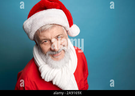 Allegro barbuto uomo invecchiato con i capelli grigi che indossa il natale costume rosso e bianco sciarpa legato sul suo collo sorridente e guardando la fotocamera. Isolato su sfondo blu. Foto Stock