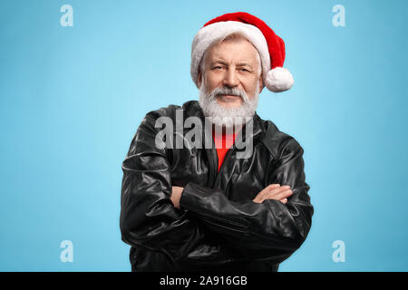 Il sambuco grey-haired maschio con barba reale e positiva espressione facciale, vestito di rosso Santa hat nero e giacca di pelle, mantenendo le mani incrociate. Uomo Senior in posa di studio con sfondo blu Foto Stock