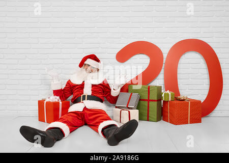 Esilarante e divertente Babbo Natale seduto sul pavimento tra splendidi multicolore di confezioni regalo e carta colorata di rosso di numeri 20. Senior uomo con i capelli grigi rilassarsi prima o dopo il lavoro di natale. Foto Stock