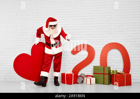 Allegro senior Saint Nicholas in tradizionale costume rosso con barba grigia e occhiali neri che mostra il pollice fino in bianco studio. Felice Natale carattere entusiasti del nuovo anno 2020 prossimamente. Foto Stock