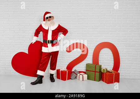 A piena lunghezza ritratto del buon vecchio Babbo Natale in nero occhiali da sole in piedi vicino al muro bianco con confezioni regalo, la carta rossa cuore e numeri di 20. Natale e Anno Nuovo concetto Foto Stock