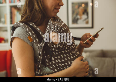 Madre con bambino in imbracatura utilizzando il telefono cellulare Foto Stock