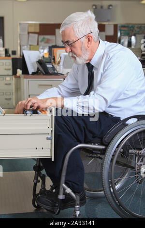 Uomo con distrofia muscolare in una sedia a rotelle archiviazione carte in il suo cassetto da ufficio Foto Stock