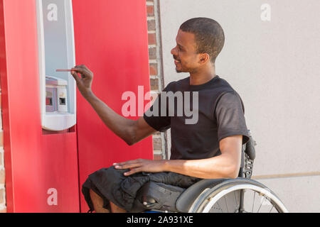 Uomo su una sedia a rotelle che ha avuto meningite spinale usando un Bancomat bancario Foto Stock