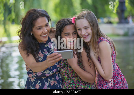 Felice madre ispanica con due figlie adolescenti con parentesi graffe che cercano in corrispondenza di messaggi di testo sul telefono cellulare in parcheggio Foto Stock