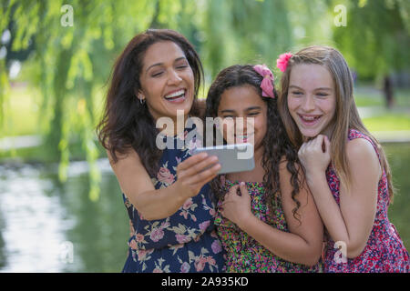 Felice madre ispanica con due figlie adolescenti con parentesi graffe che cercano in corrispondenza di messaggi di testo sul telefono cellulare in parcheggio Foto Stock