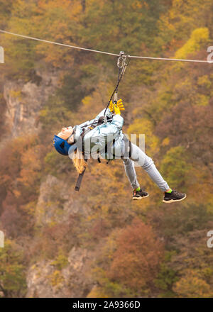 Una ragazza adolescente vola su un zipline contro lo sfondo delle montagne coperte di bosco d'autunno. Foto Stock