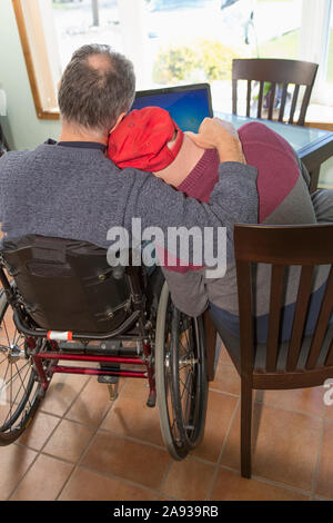 Giovane uomo con sindrome di Down abbracciando suo padre in sedia a rotelle Con lesione del midollo spinale a casa Foto Stock