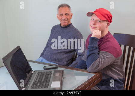 Felice giovane uomo con la sindrome di Down e suo padre dentro Sedia a rotelle con lesioni al midollo spinale utilizzando un computer portatile a casa Foto Stock