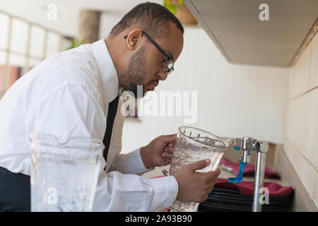 L'uomo afroamericano con la sindrome di Down come un cameriere che riempie caraffa d'acqua in cucina Foto Stock