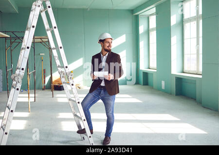 Caposquadra uomo barbuto in un casco bianco in una camera presso un sito in costruzione Foto Stock