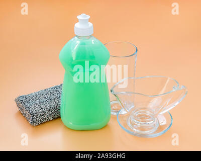 Piatto verde del liquido di lavaggio in una plastica trasparente bottiglia, argento spugna di schiuma e vetreria su un pastello sfondo arancione. Detergente da cucina. Foto Stock