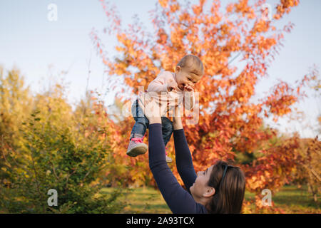 Vista laterale di felice giovane madre holding Baby girl nelle sue mani e guardando a Lei con amore. Autunno photosession Foto Stock