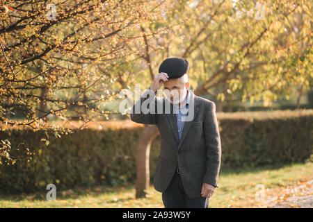 Bel uomo anziano mettendo su una giacca grigia e cappello nero. Old grey-haired uomo barbuto a piedi nel parco d'autunno. Sfondo giallo Foto Stock