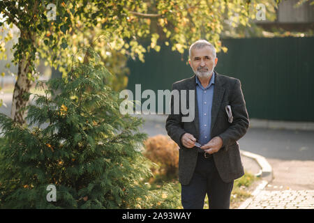 Bello il senior uomo a piedi attraverso il parco con il giornale in mano. Il nonno barbuto di prima mattina a piedi nel parco Foto Stock