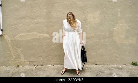 Giovane donna bionda che indossa lungo abito bianco guardando in giù a terra di fronte grungy parete in stucco Foto Stock