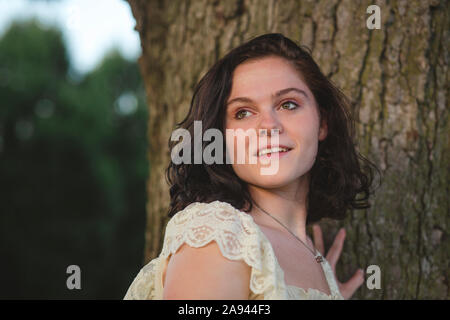 Ritratto di un sorridente giovane donna in piedi contro un albero al tramonto Foto Stock