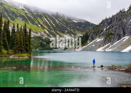 Una donna si blocca sul lato di un lago alpino con il suo cane e prende in vista di costa le montagne intorno a Pemberton, British Columbia, in una piovosa giornata d'estate. Foto Stock