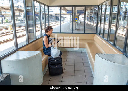 Passeggeri donne che usano il suo smartphone in attesa di un treno con valigia nella stazione ferroviaria FFS di Basilea; Basilea, Basilea Città, Svizzera Foto Stock