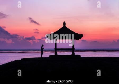 Profilo di uomo in piedi accanto alla pagoda di mare sulla spiaggia di Sanur, Bali, Indonesia a sunrise. Mare di lavanda in background; nuvole e colorato del cielo. Foto Stock