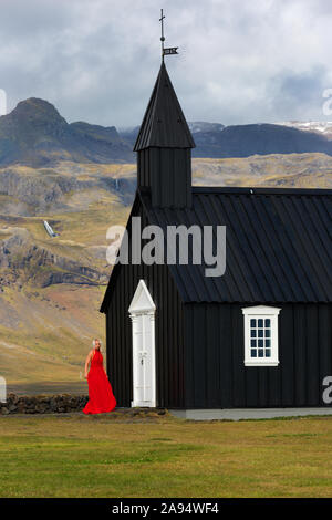 Signora in abito rosso Budir chiesa un nero struttura in legno dal 1847 seduto sulla penisola Snaefellsnese , Islanda Foto Stock
