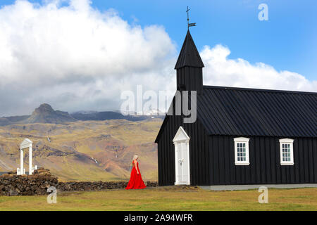 Signora in abito rosso Budir chiesa un nero struttura in legno dal 1847 seduto sulla penisola Snaefellsnese , Islanda Foto Stock