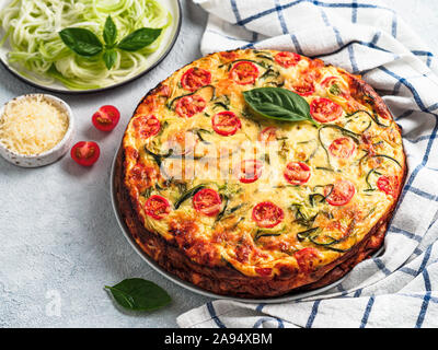 Tortino di zucchine e gli ingredienti su sfondo grigio, copia spazio a sinistra. Deliziosa torta salata con zucchine, pomodori, erbe e formaggio. Idea e ricetta per cottura sana e molto per il raccolto di zucchine Foto Stock