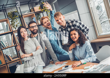 Startupers lavorando insieme a Office abbracciando permanente guardando la telecamera allegro Foto Stock