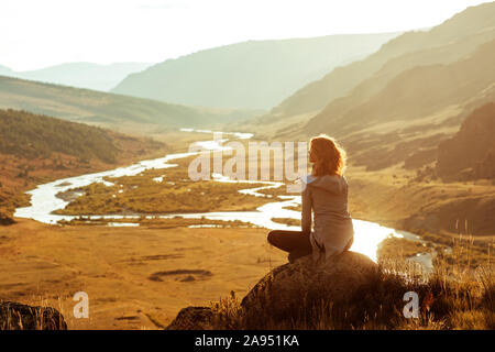 Slim ragazza siede sul big rock e gode della vista del tramonto montagne e il fiume Foto Stock