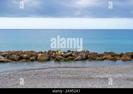 Un uomo non identificabili con benne e le aste pesche sugli scogli lungo la costa di Ventimiglia, Italia, sulla Riviera Italiana. Foto Stock