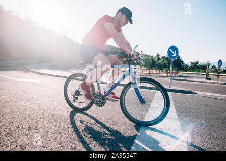 Montare maschio ciclista biker in sella alla sua moto ciclo su una strada asfaltata al tramonto mentre il sole tramonta attraverso la sua ruota. Foto Stock