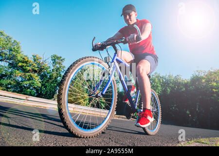 Montare maschio ciclista biker in sella alla sua moto ciclo su una strada asfaltata al tramonto mentre il sole tramonta attraverso la sua ruota. Foto Stock