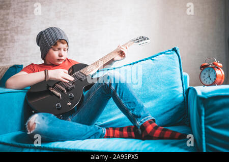 I capelli rossi boy in jeans e un cappuccio seduti sul divano esegue un esercizio sulla riproduzione di un nero chitarra elettrica - rock e rock-roll style Foto Stock
