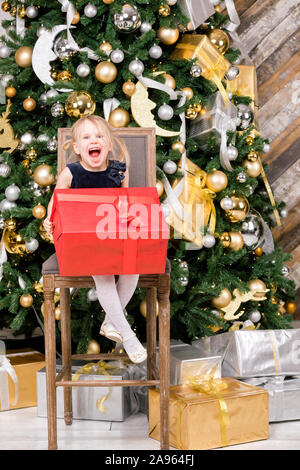 Ritratto di gioiosa elementari di età ragazza indossa vestito blu seduti su alti sedia accanto all'albero di Natale azienda big golden avvolto confezione regalo felice t Foto Stock