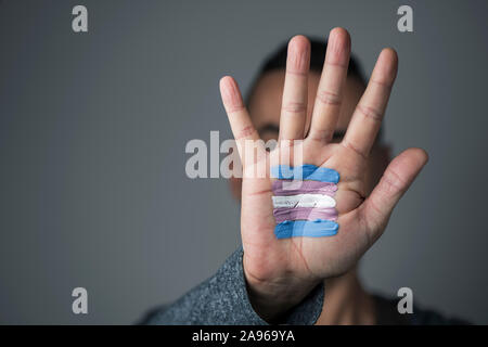 Primo piano del palmo della mano di un giovane uomo caucasico con un transgender bandiera dipinta in esso, di fronte al suo o il suo volto Foto Stock