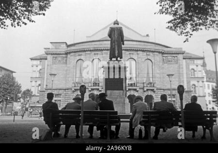 Mainzer sitzen auf Bänken auf dem Gutenbergplatz und blicken auf das Gutenbergdenkmal und das Stadttheater in Mainz, Deutschland 1961. Abitanti di Magonza seduti sui banchi sulla Gutenbergplatz square, rivolto verso la parte posteriore del monumento di Gutenberg e la città del teatro, Germania 1961. Foto Stock