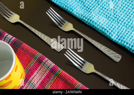 Tre forchette argentate tra tovaglioli blu e multicolore con coppa gialla sullo sfondo scuro riflettente. Linee e posate blu e colorate. Foto Stock
