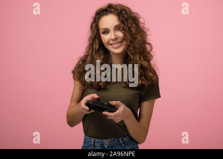 Ritratto di Pretty girl geek con capelli ricci azienda gamepad e sorriso isolato su sfondo rosa Foto Stock