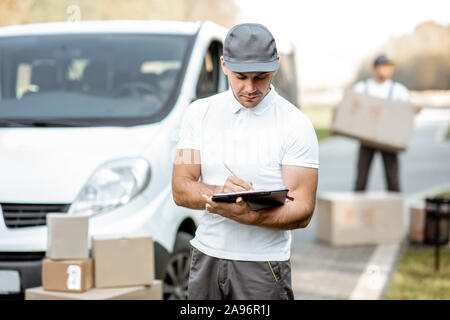 Ritratto di consegna uomo in uniforme in piedi con documenti vicino il cargo FURGONE veicolo, collega scarica pacchi sullo sfondo Foto Stock