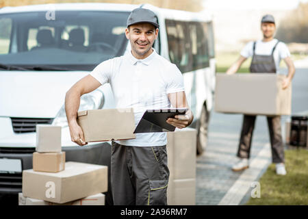 Ritratto di consegna uomo in uniforme in piedi con documenti vicino il cargo FURGONE veicolo, collega scarica pacchi sullo sfondo Foto Stock