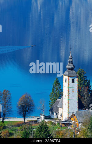 Splendida vista della chiesa cattolica di Giovanni Battista presso il lago di Bohinj in autunno e pescatore solitario in canoa su una mattina di sole in Slovenia. Foto Stock