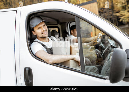 Allegra compagnia di consegna dipendenti in uniforme il carico di guida il veicolo, la consegna delle merci ai clienti, bello corriere con pacco sul sedile del passeggero Foto Stock