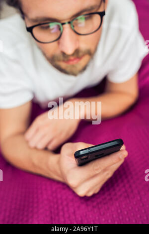 Foto di un piuttosto grave giovane uomo bello interagire con il suo dispositivo mobile mentre giaceva a letto Foto Stock