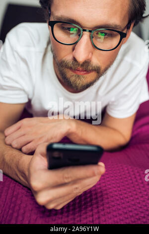 Foto di un piuttosto grave giovane uomo bello interagire con il suo dispositivo mobile mentre giaceva a letto Foto Stock