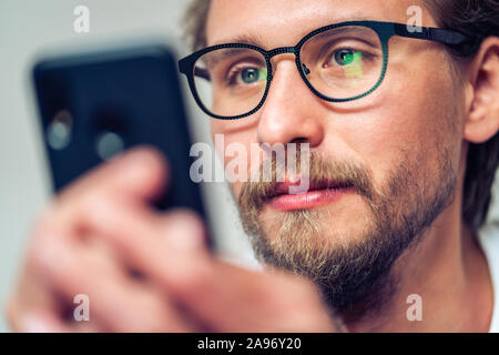 Piuttosto giovane serio con gli occhiali è sdraiato sul letto mentre guardando al suo telefono cellulare Foto Stock