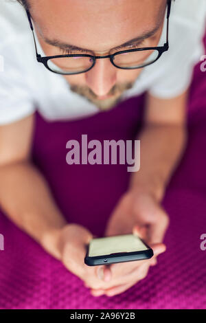 Foto di un piuttosto grave giovane uomo bello interagire con il suo dispositivo mobile mentre giaceva a letto Foto Stock