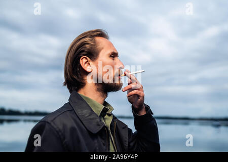 Foto di un giovane uomo barbuto di fumare una sigaretta. Il lago e le nuvole in background. Foto Stock